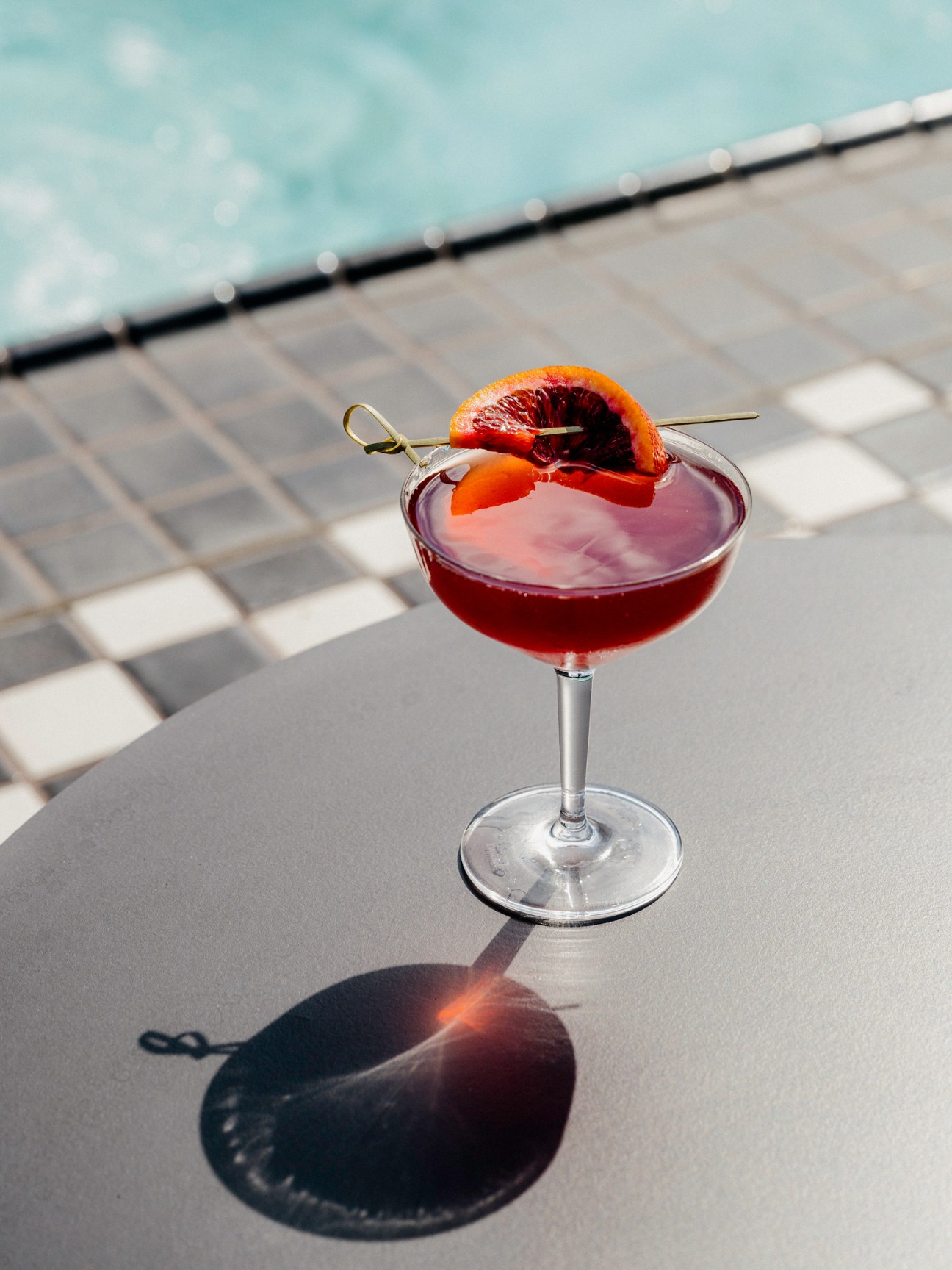 Dark cocktail in coupe glass with orange slice garnish and cocktail pick on outdoor table