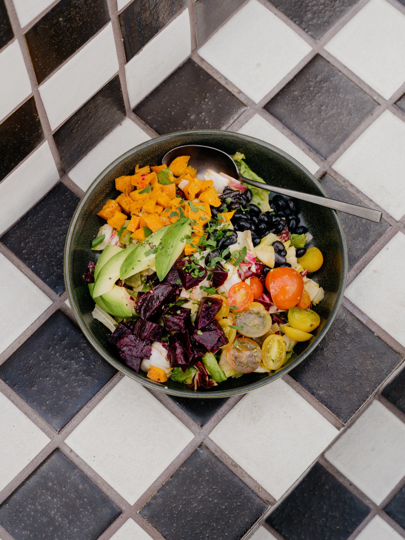 Colorful salad bowl with avocado, black beans, cherry tomatoes, and mixed greens on checkered tile surface