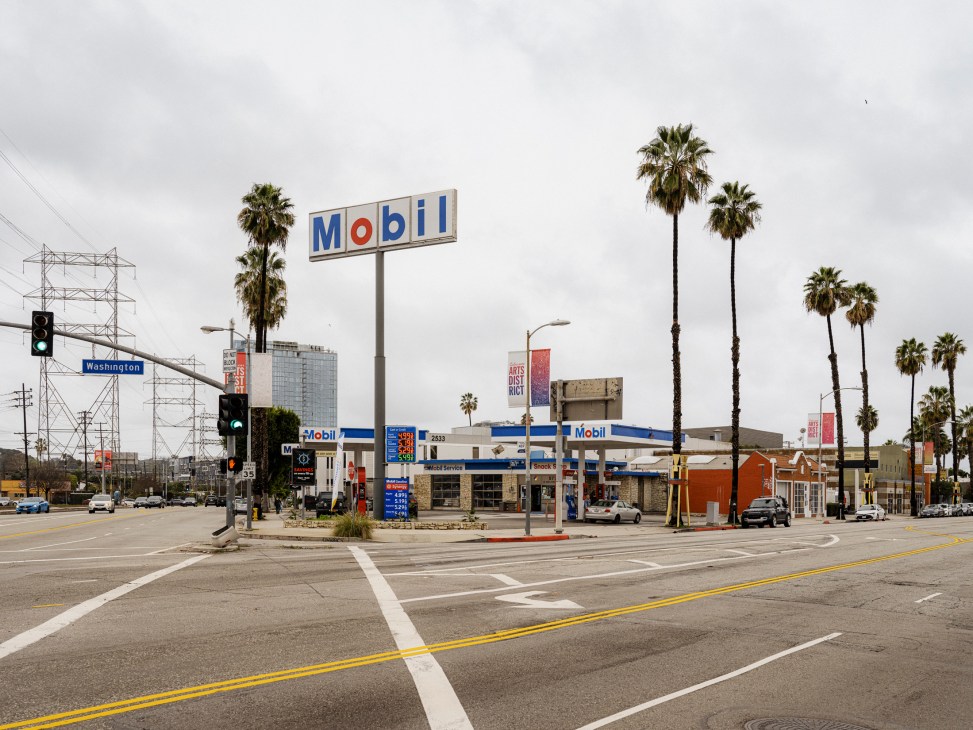A vintage Mobil gas station with white and blue signage surrounded by palm trees on a wide street intersection