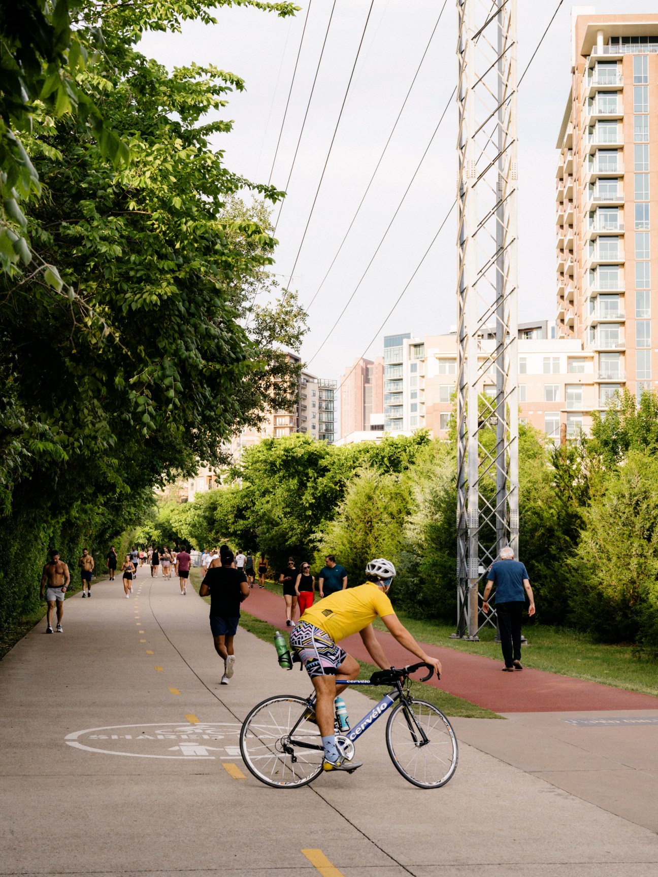 A cyclist in yellow rides along the Katy Trail with downtown Dallas apartments and power lines visible in background