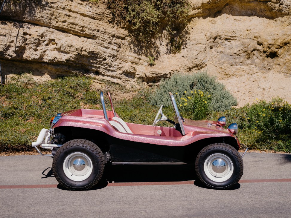 Red and black vintage-style Meyers Manx dune buggy parked on road with rocky cliff background