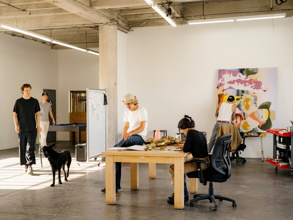 Open artist studio space with people working at tables, exposed ceiling, white walls, and colorful artwork displayed