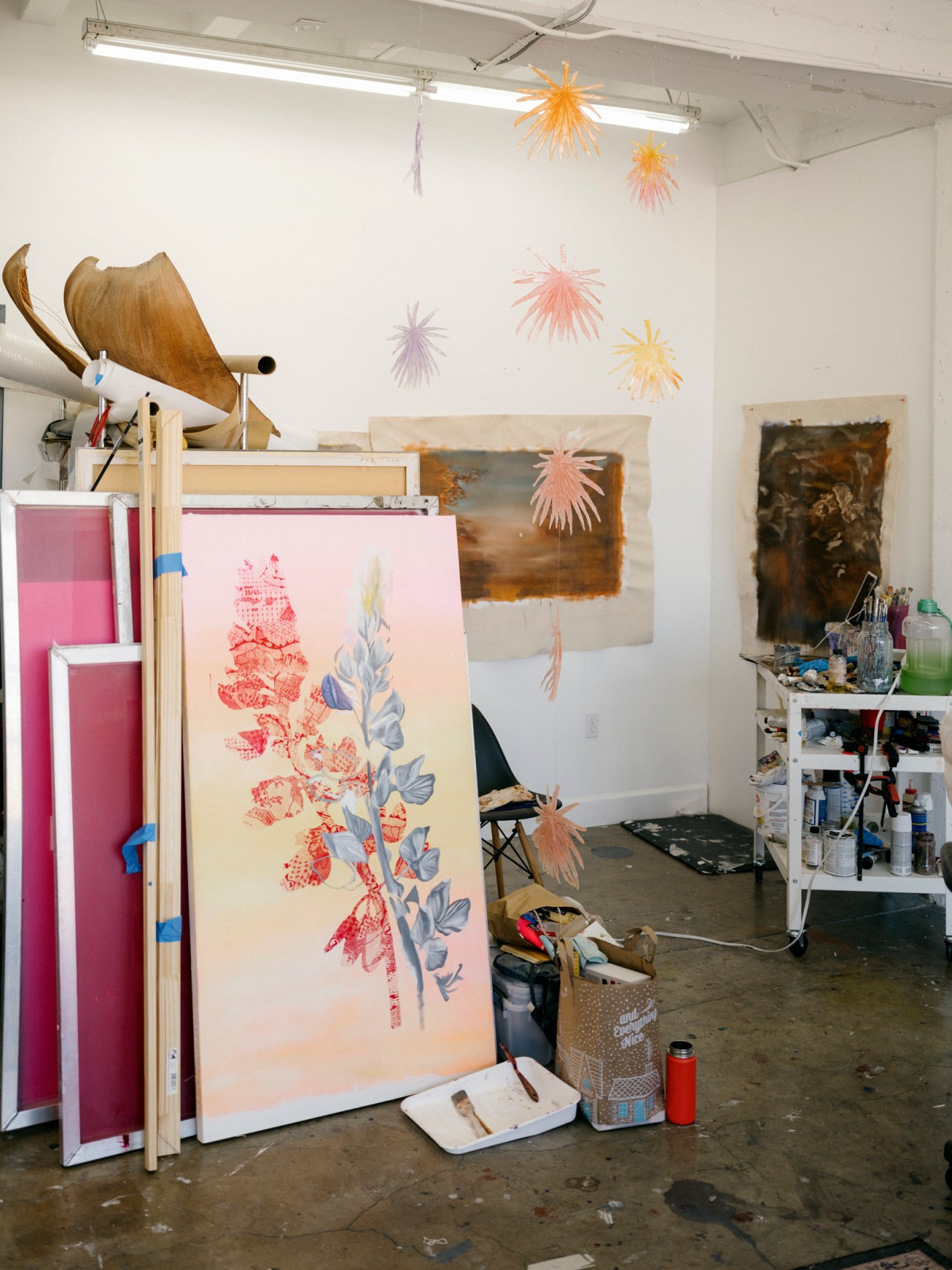Corner of an artist studio showing easel with botanical artwork, hanging paper decorations, and art supplies