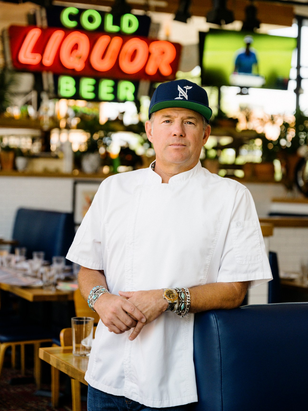 Chef Nick Badovinus in white chef coat and baseball cap standing in restaurant with neon signage