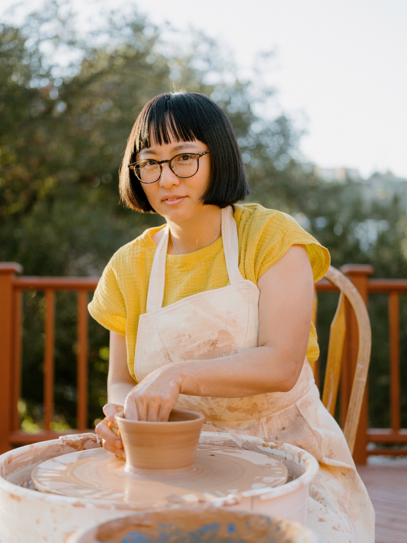 Poppy Shibamoto working with clay at a pottery wheel, wearing glasses and a yellow shirt with white apron
