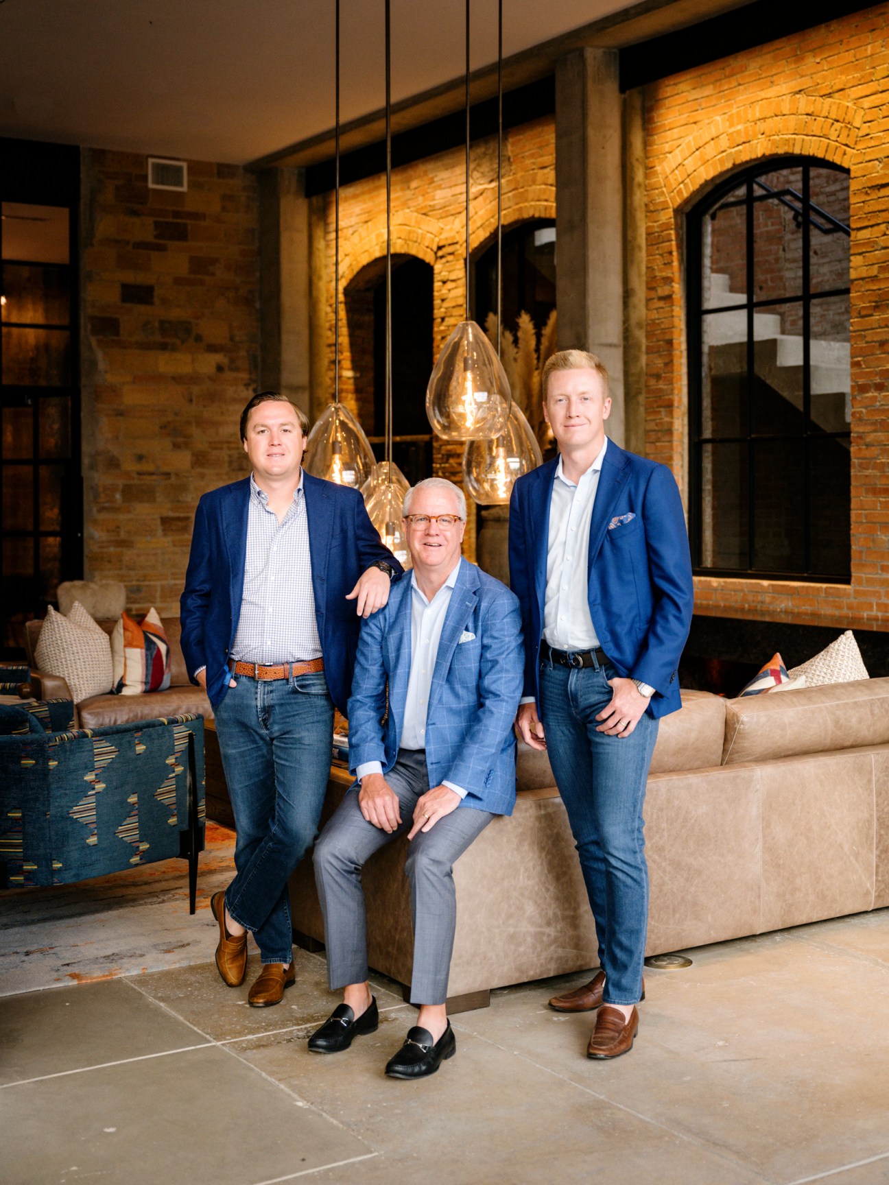 Three men in blue blazers pose in a modern interior with exposed brick walls and pendant lighting