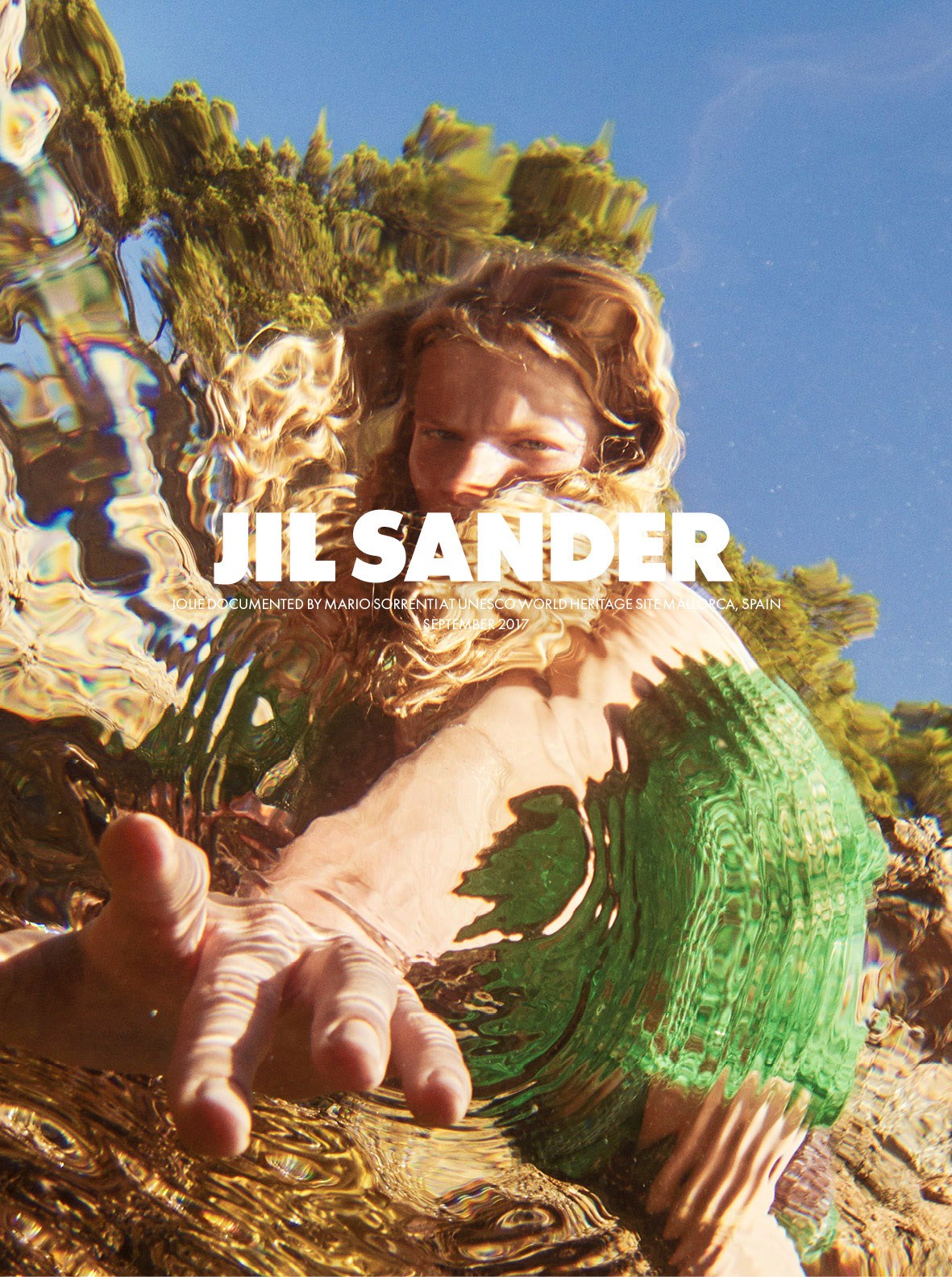 Model in white clothing among golden sculptural elements and green foliage under blue sky for Jil Sander campaign