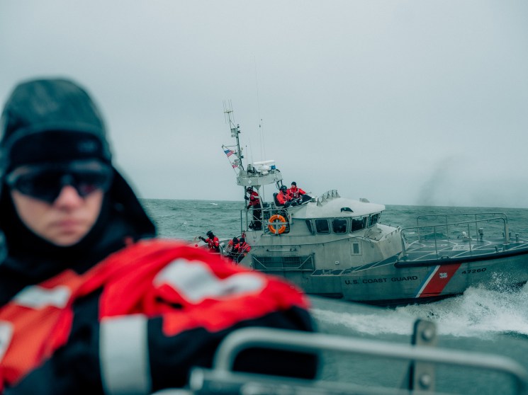 Coast Guard member in protective gear on boat with rescue vessel approaching in rough seas