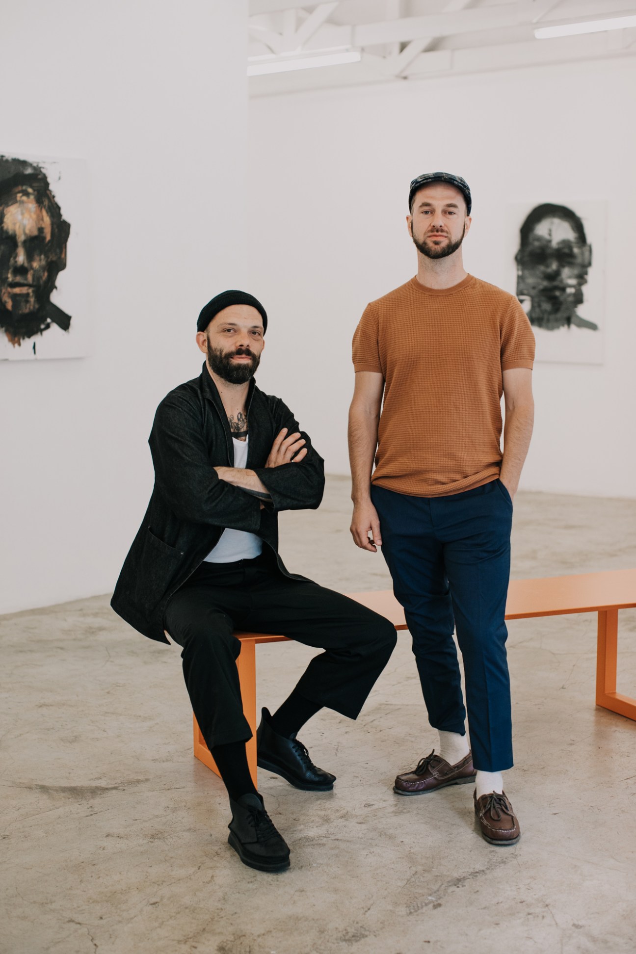 Two men standing in a white-walled gallery space with artwork visible on the walls behind them