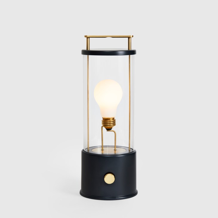 The Muse portable lantern by Tala with black cylindrical base, brass frame, and illuminated bulb inside glass cylinder