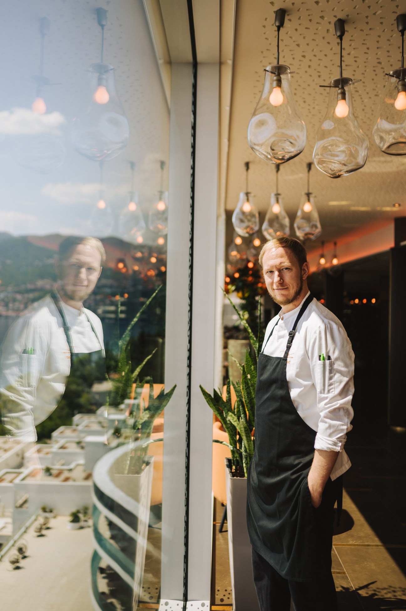 Chef Deni Srdoc in white chef's jacket and black apron standing in a modern restaurant interior