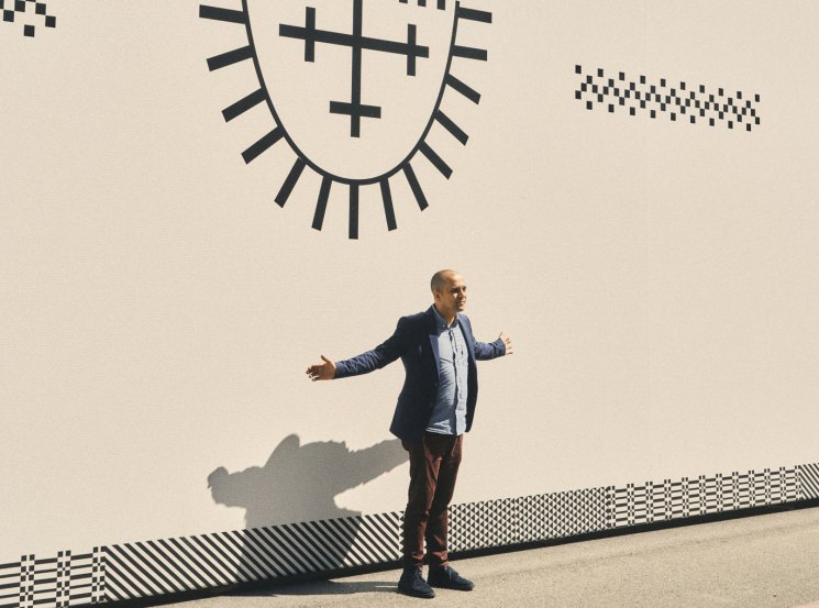 Man in casual attire standing against white wall with large artistic mural featuring geometric patterns