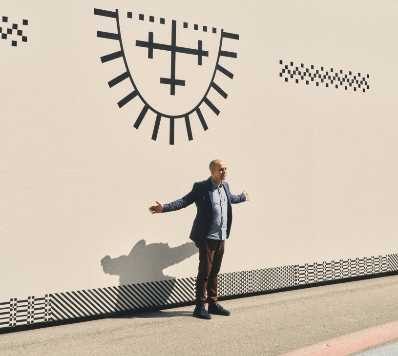 Man in casual attire standing against white wall with large artistic mural featuring geometric patterns