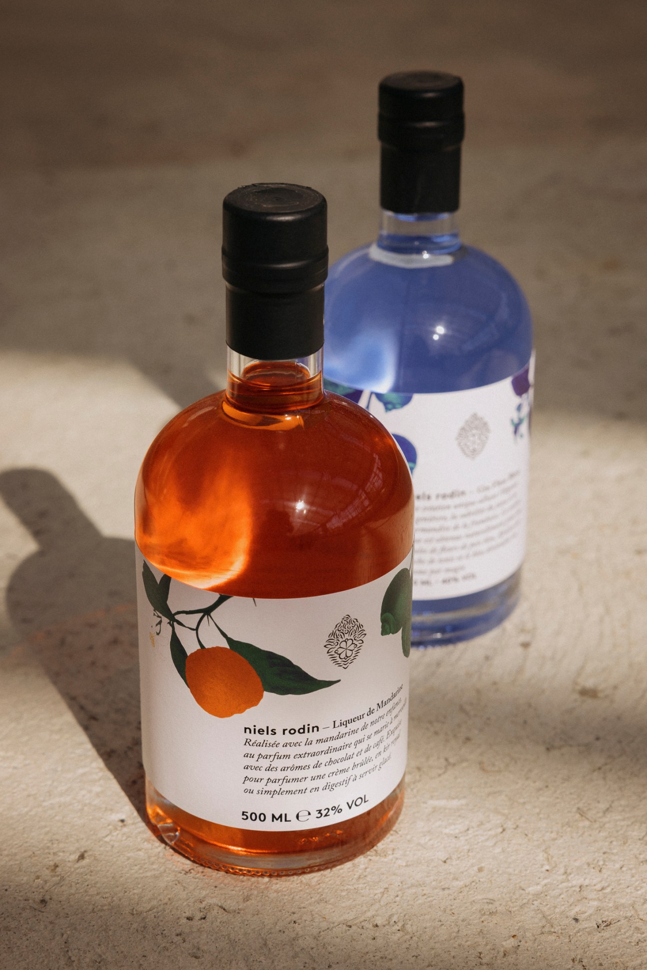 Two bottles of spirits: Niels Rodin's Liqueur de Mandarine with amber liquid and mandarin leaf label, and Gin Fleurs Bleus with blue liquid.