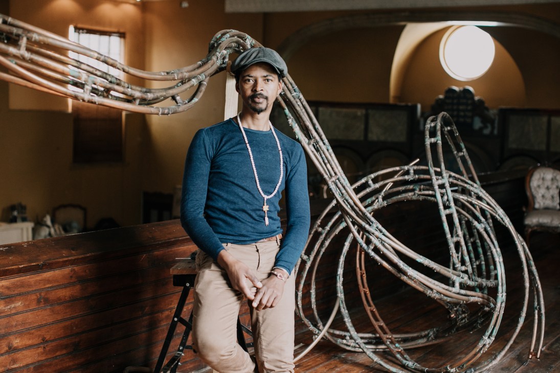 Nicholas Hlobo sits wearing a blue shirt and head wrap surrounded by curved metal sculptural elements in his studio