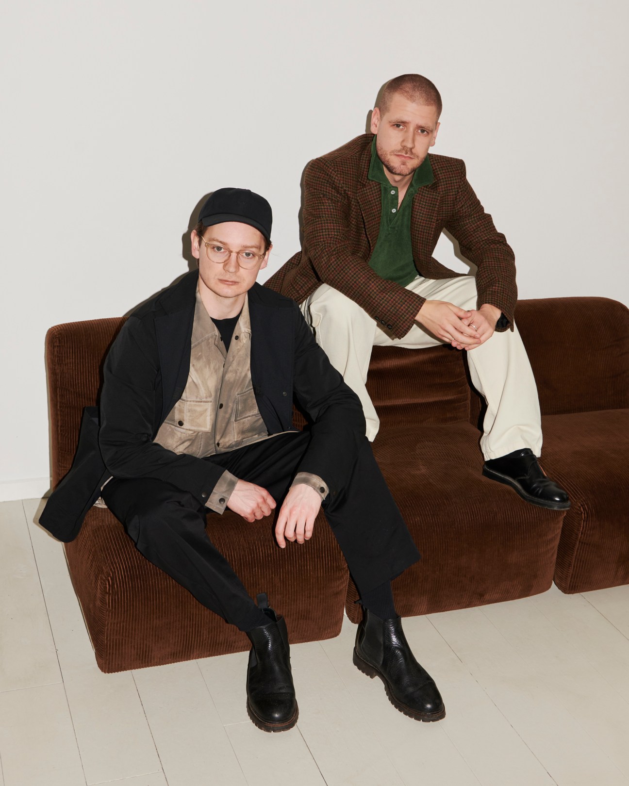 Two men sitting on a brown couch, one wearing black outfit with beanie, the other in brown checked blazer