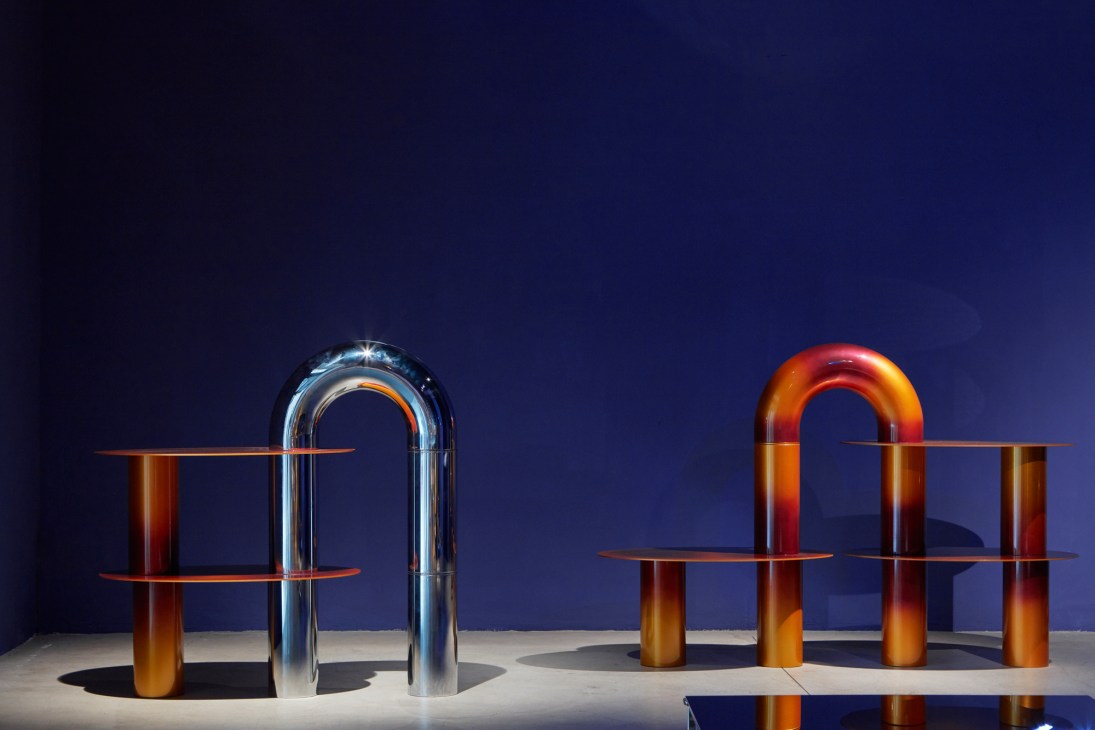 Modern sculptural furniture with curved glass and brass elements against a deep blue backdrop.