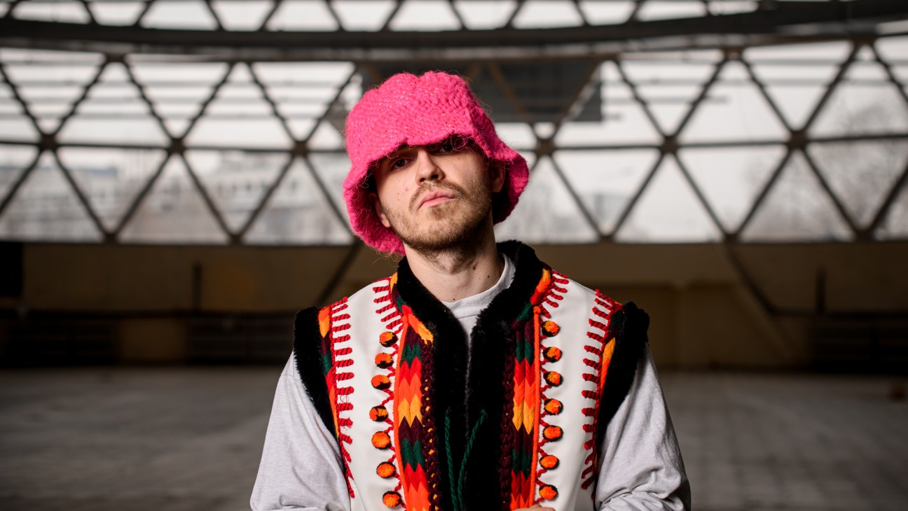 Young man wearing bright pink knit bucket hat and colorful traditional vest standing in urban setting