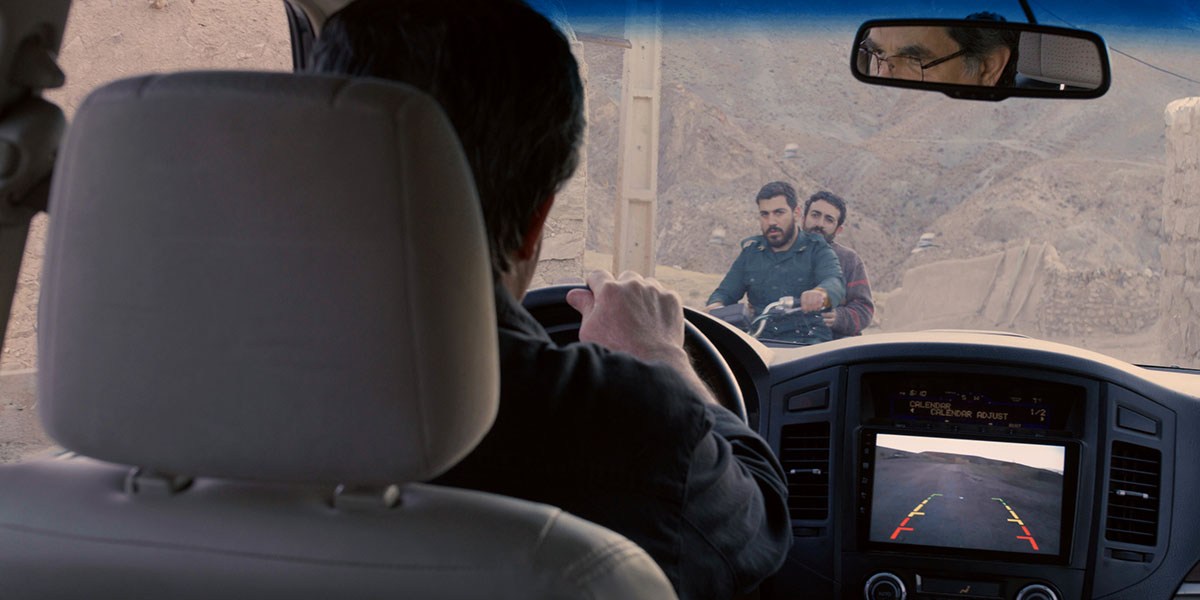 View from inside a car showing two men on a motorcycle outside in an arid landscape