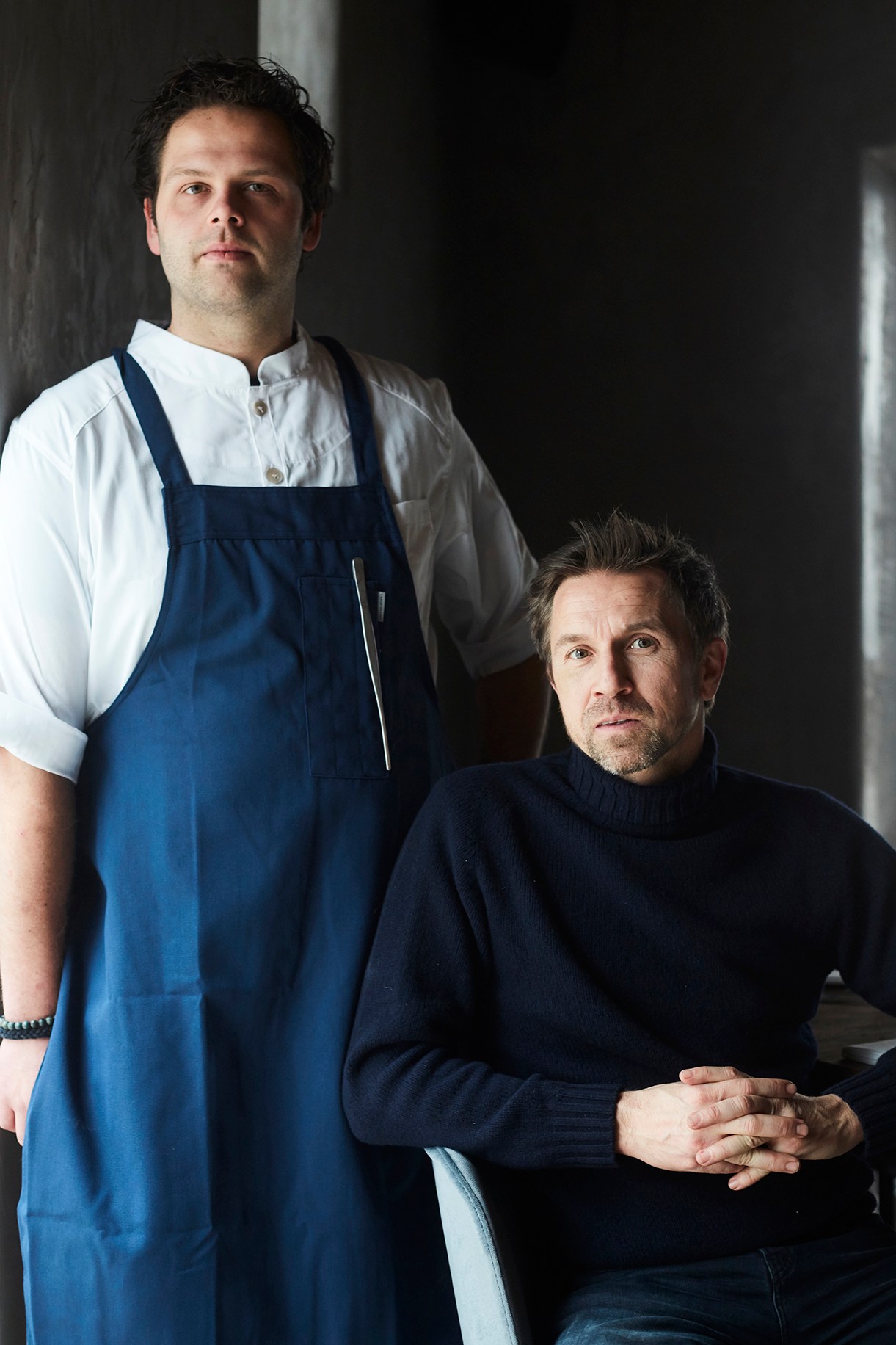 Two men posing for portrait, one standing wearing chef's apron and white shirt, one seated wearing black turtleneck