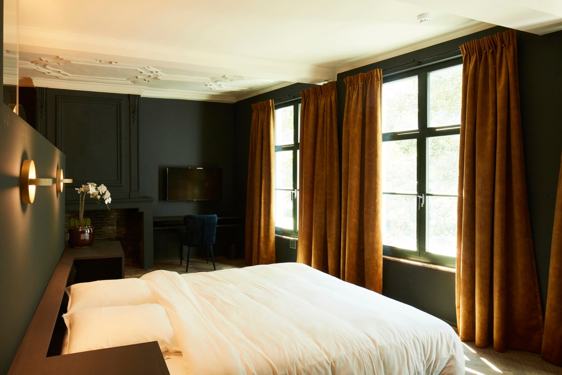 Modern hotel bedroom with dark green walls, white bedding, orange curtains and French doors