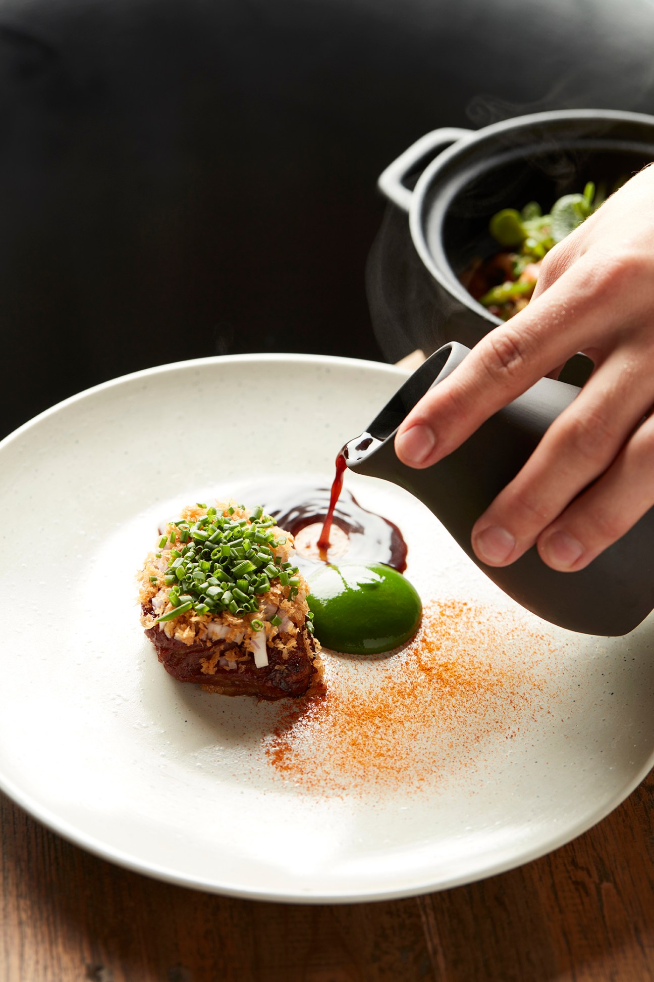 Hand drizzling dark sauce from small bottle onto plated dessert with green garnish and powdered spice