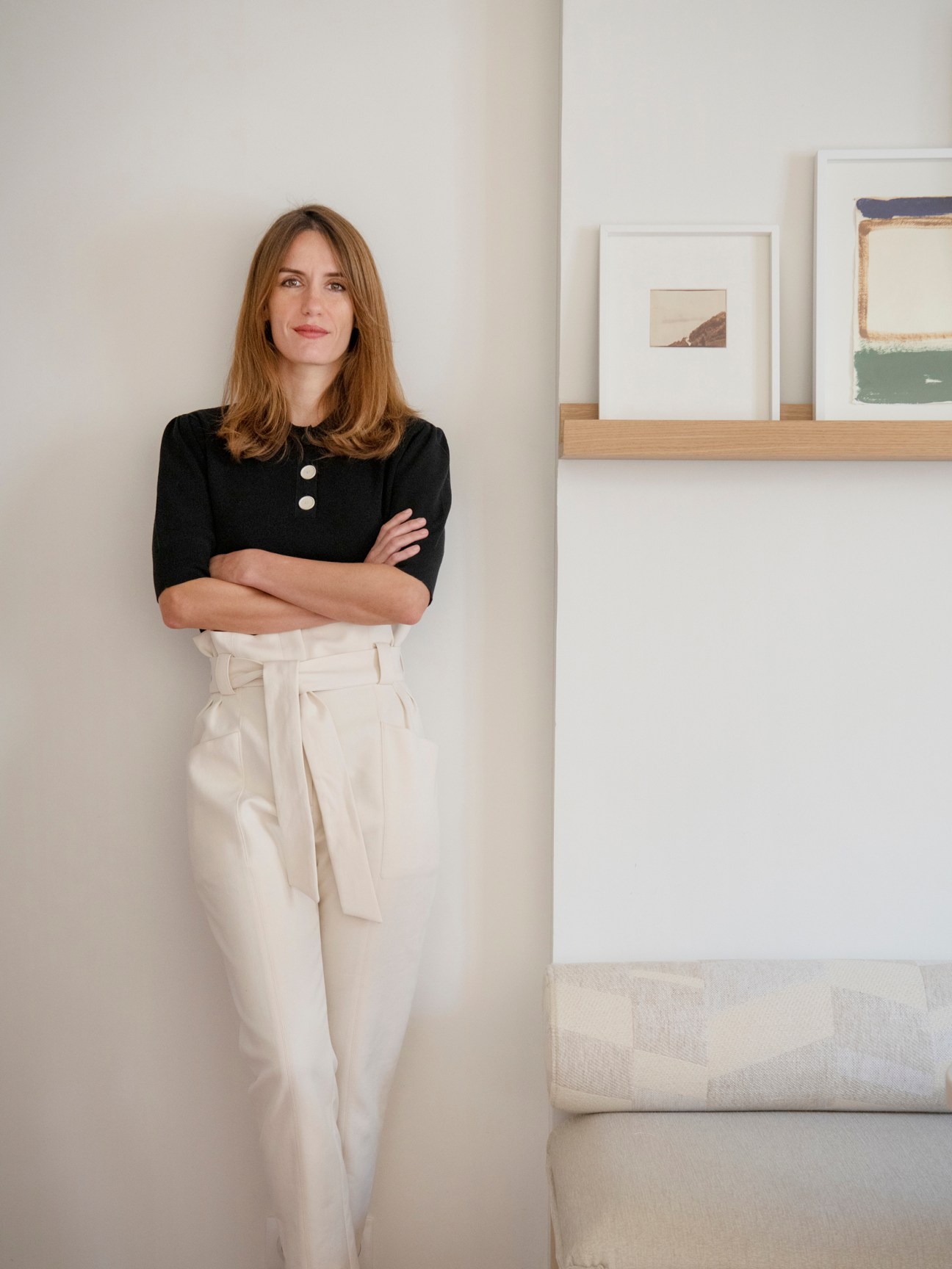 Woman in black top and cream trousers standing with crossed arms in minimalist interior