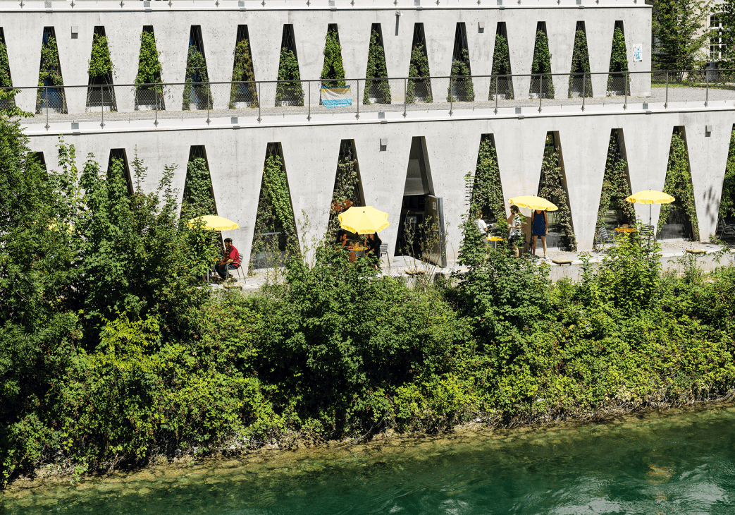 Tiered concrete structure with triangular openings overlooking water, people with yellow umbrellas in alcoves