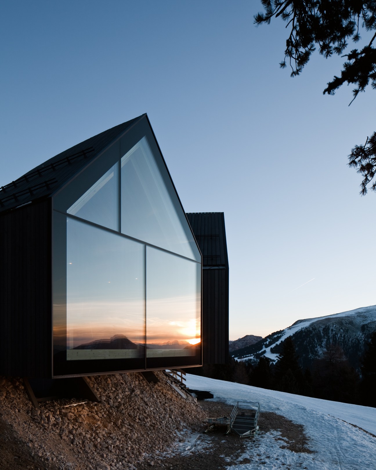 Modern glass mountain hut with triangular windows reflecting sunset over snowy alpine landscape