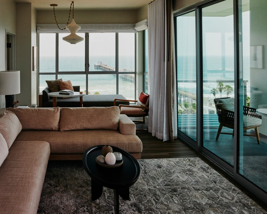 Hotel living room with beige sectional sofa, black coffee table, and large windows overlooking ocean and pier
