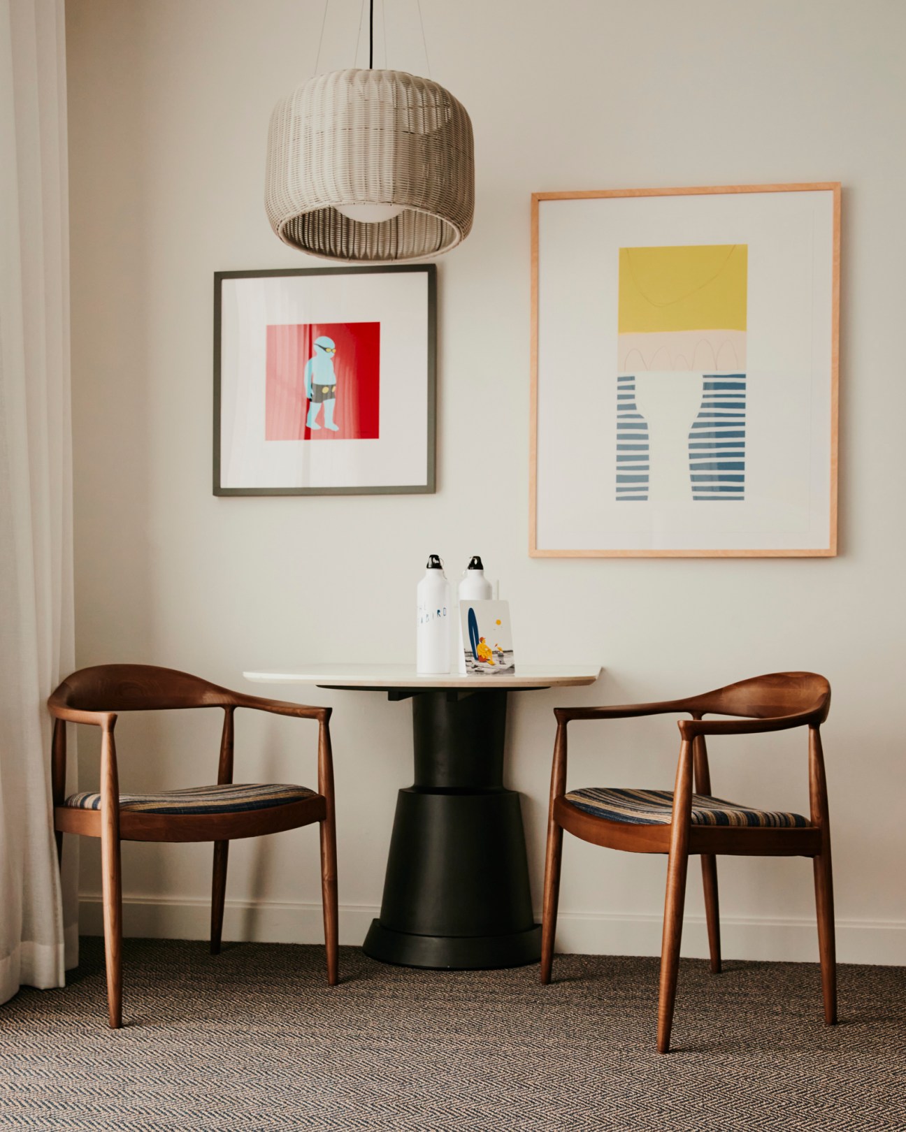 Modern dining area with two wooden chairs, round table, woven pendant light, and two framed artworks on white wall