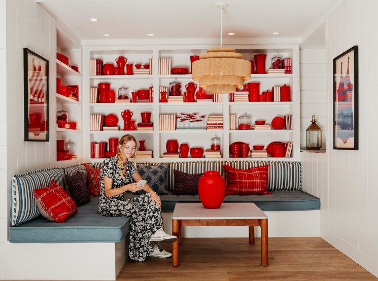 Woman in patterned dress sitting on built-in banquette with striped pillows beneath shelving displaying red pottery and books