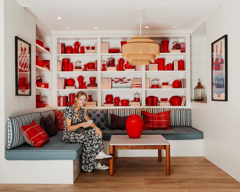 Woman in patterned dress sitting on built-in banquette with striped pillows beneath shelving displaying red pottery and books