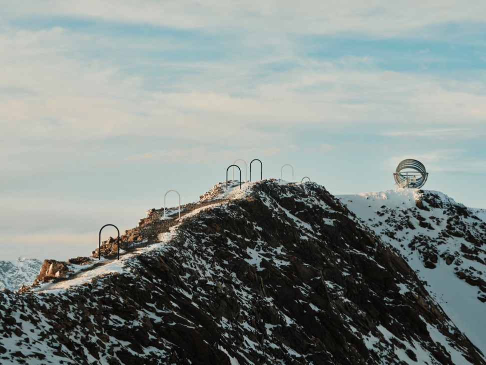 Metal archways mark a snowy mountain ridge leading to Olafur Eliasson's spherical sculpture on an Alpine peak.