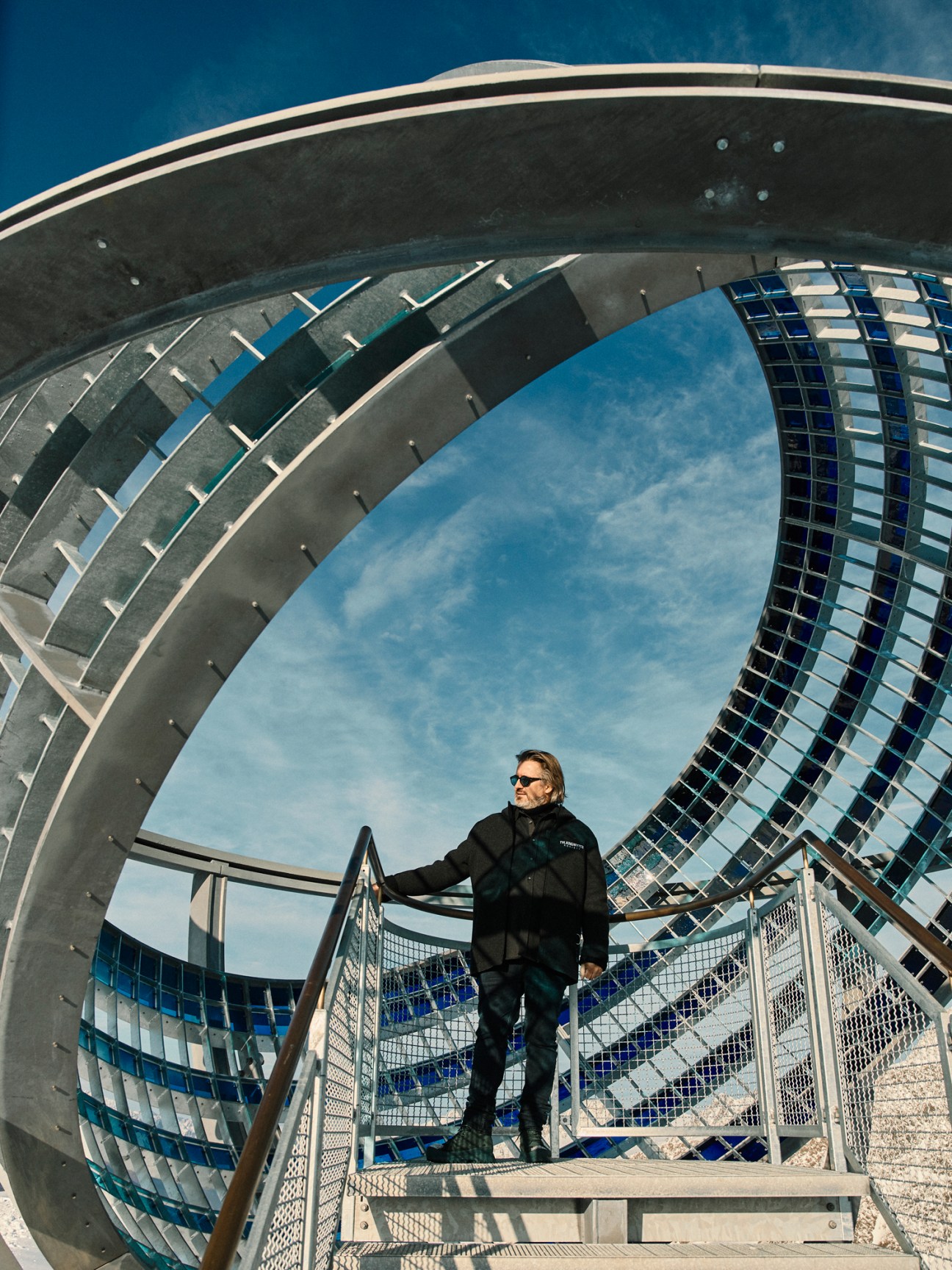Olafur Eliasson stands inside the Our Glacial Perspectives sculpture, a spherical steel and glass structure with blue-tinted panes that frame the sky.