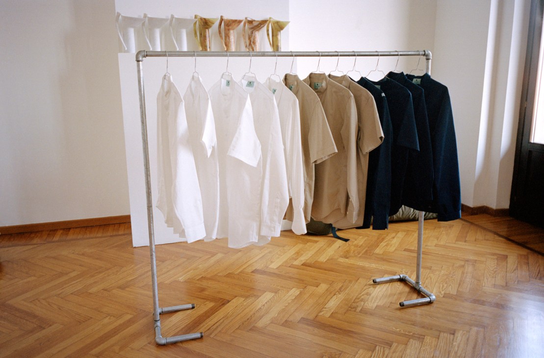 Clothing display at Older studio showing white, cream, tan and black garments hung on a metal rack in a minimalist white gallery space.