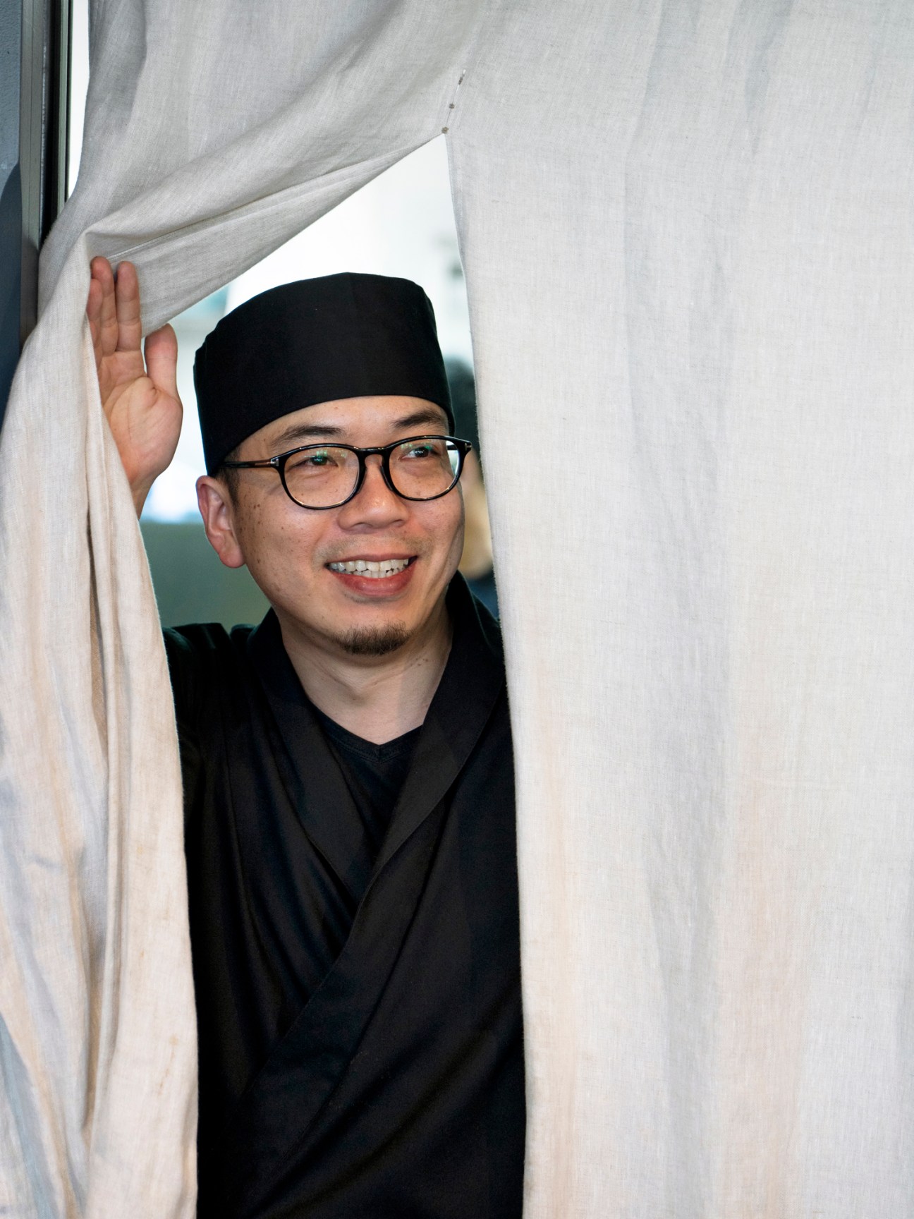 Chef peering through sheer curtains at Ooki Pavilion restaurant, wearing glasses and black chef's attire