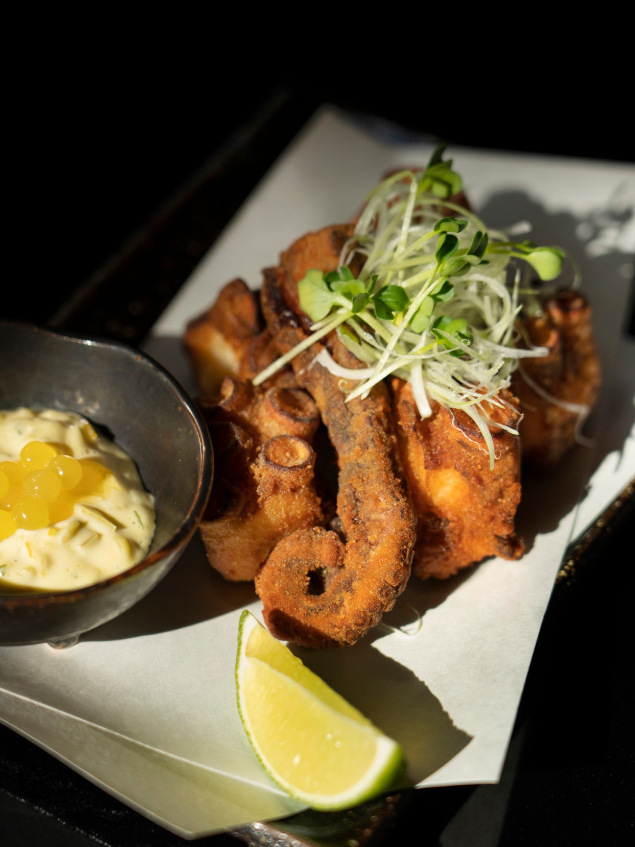 Fried octopus served on a white plate with microgreens, lime wedge, and dipping sauce.