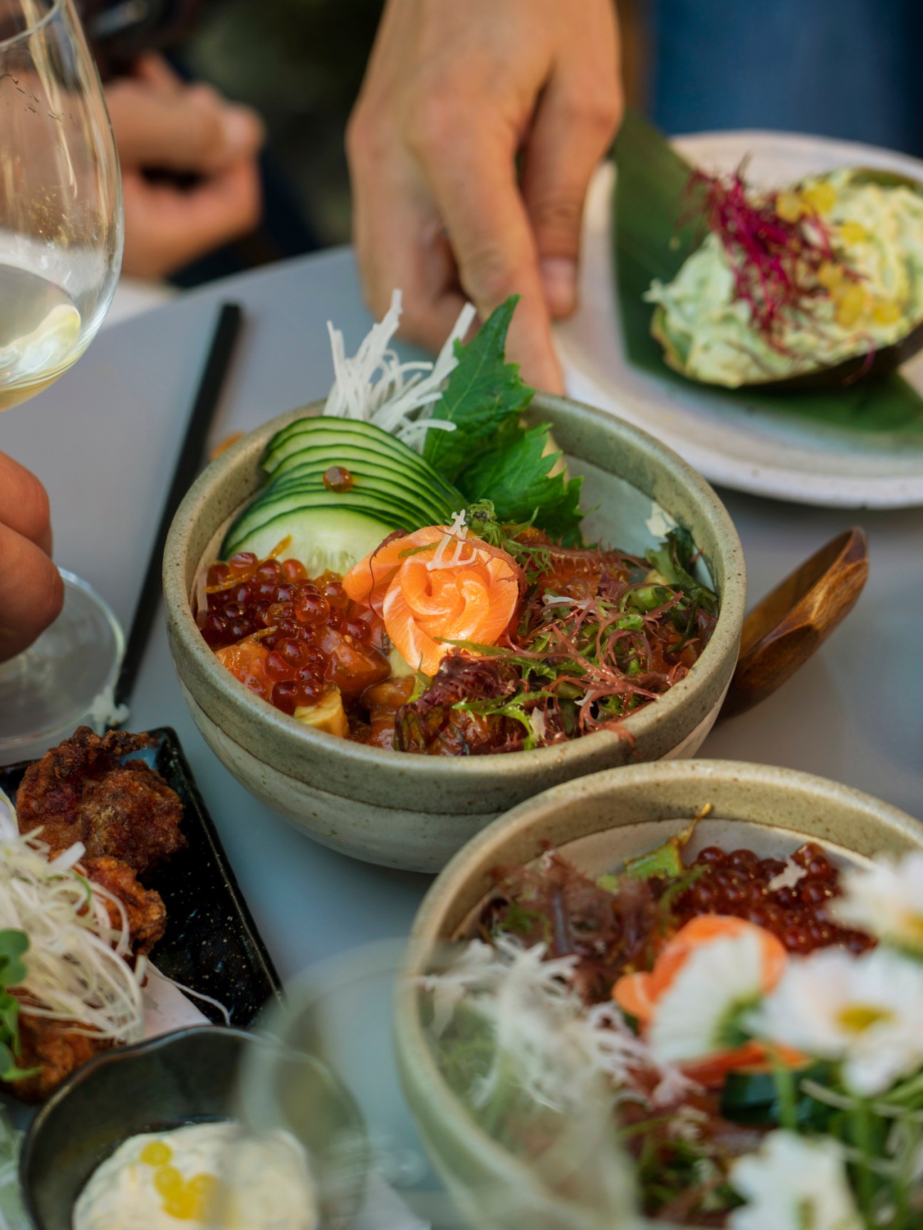A hand reaching toward a ceramic bowl of Japanese sashimi and sushi rice with sliced cucumber, salmon roe, and garnishes at Ooki Pavilion restaurant.