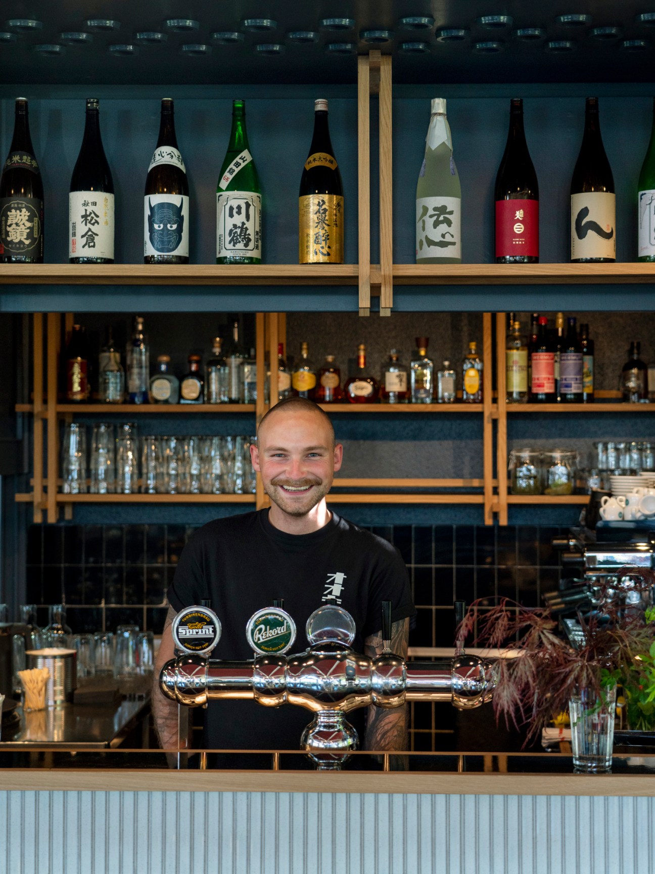 Bartender Robin smiling behind the bar at Ooki Pavilion with saké bottles and shelves of spirits.