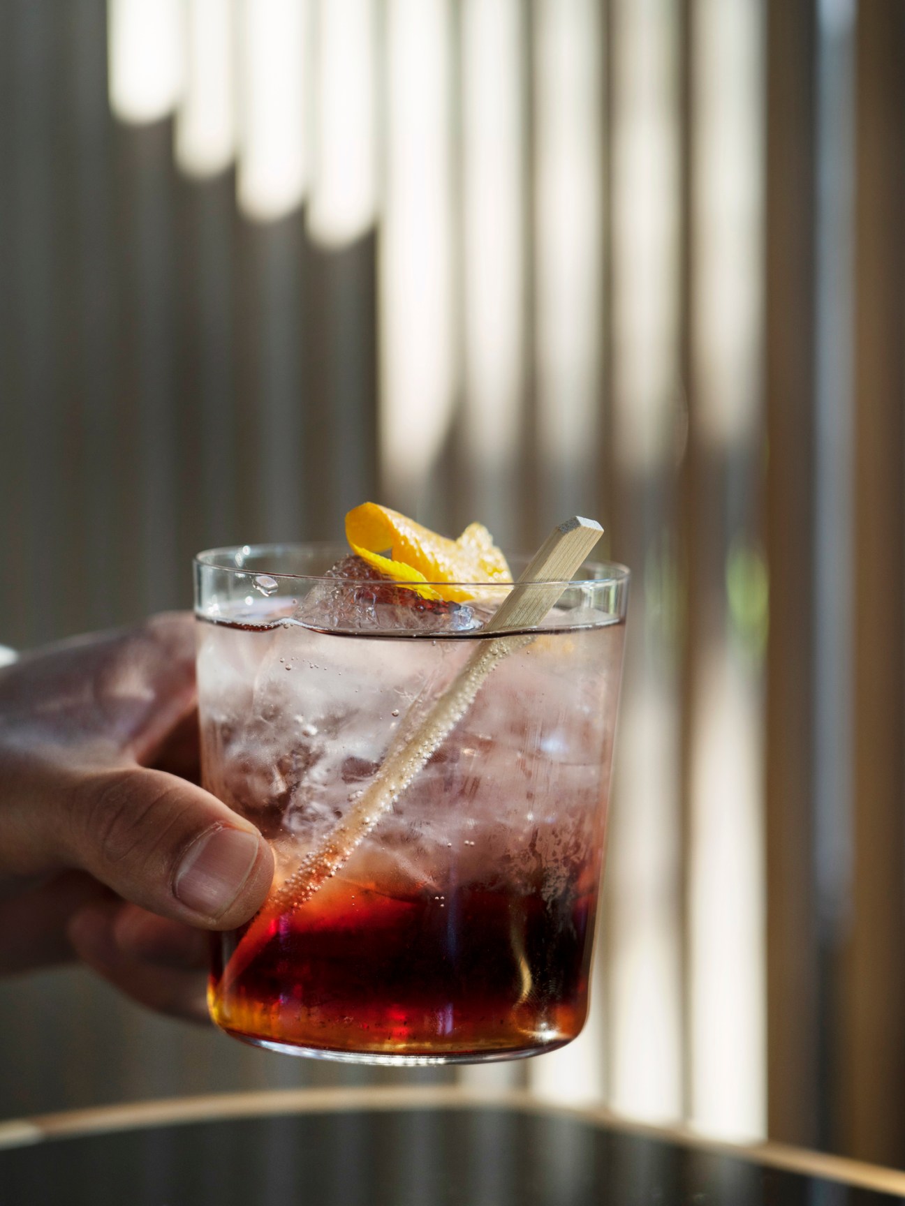 Hand holding a glass of umericano cocktail with lemon twist and wooden stir stick, with blurred building exterior in background.