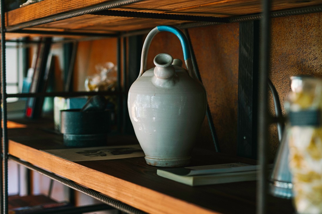 Gray ceramic jug with lid and handle sits on wooden shelf next to books