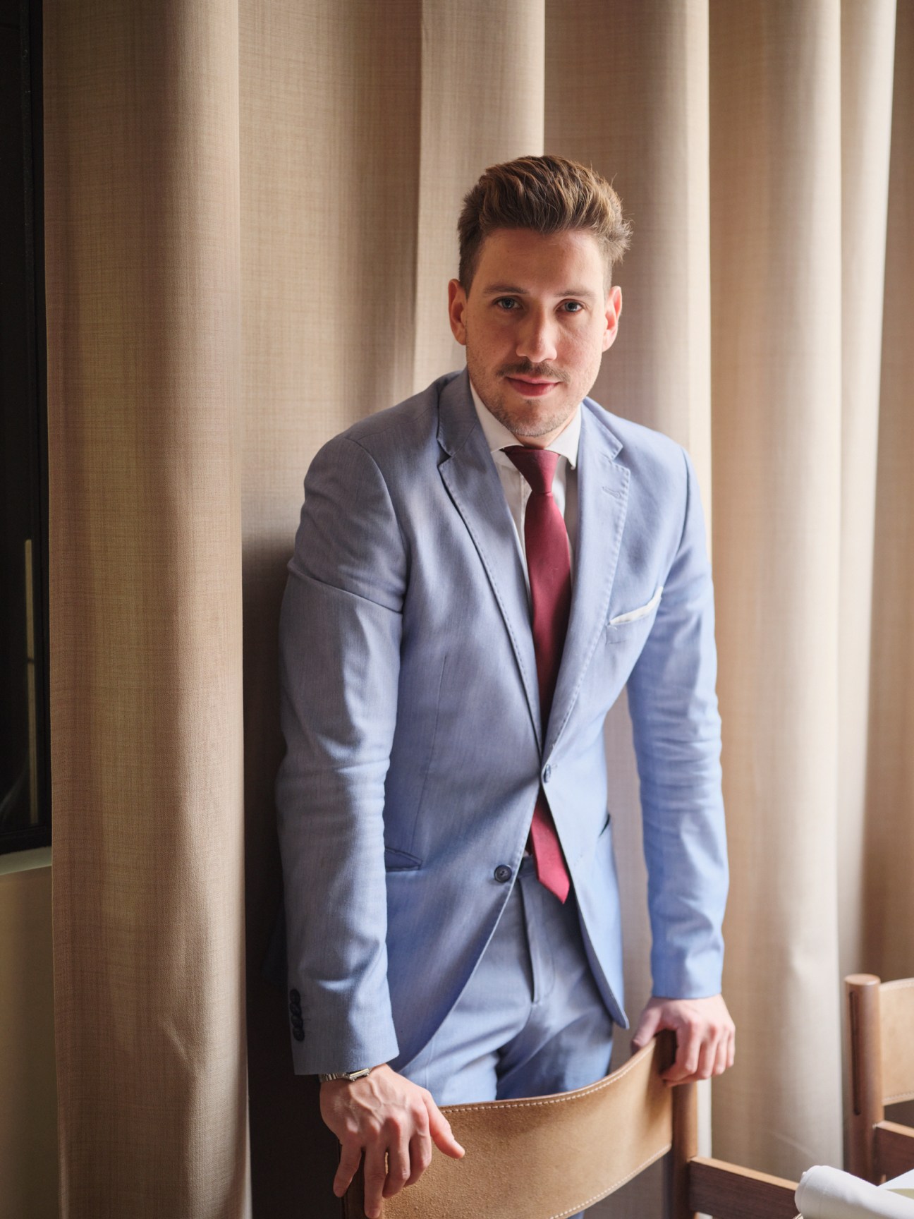 Man in light blue blazer and burgundy tie standing against cream curtains.