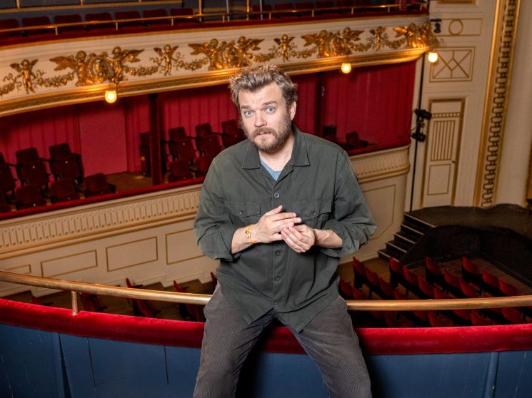 Pilou Asbaek sits on a theater balcony railing in an ornate theater with red velvet seating