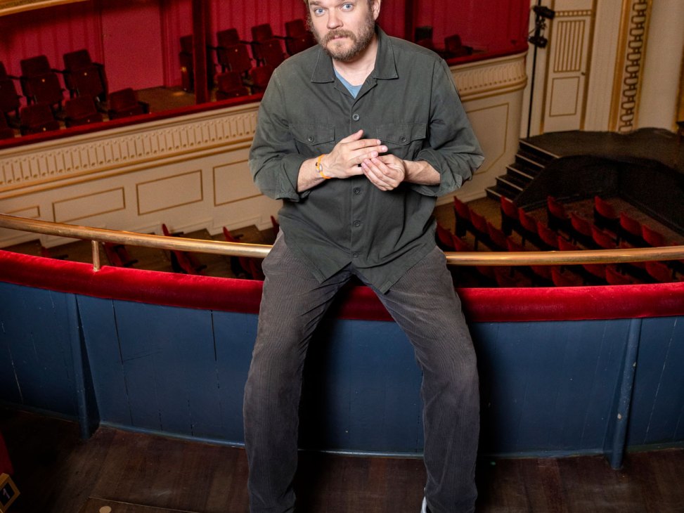 Pilou Asbaek sits on a theater balcony railing in an ornate theater with red velvet seating