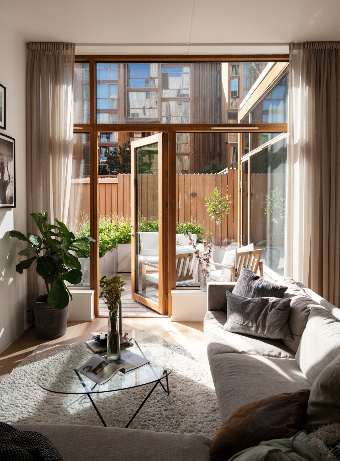 Minimalist apartment interior with grey sofa, potted plant, and floor-to-ceiling wooden doors opening onto a planted balcony with building views.