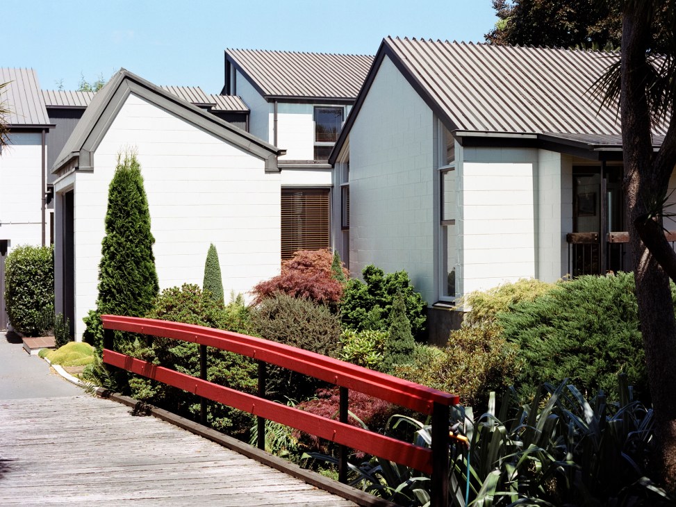 Modern house exterior with white concrete block walls, steep pitched roof, and landscaped garden with red timber bridge