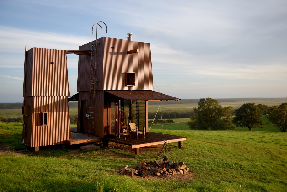 Modern timber cabin with corrugated metal cladding and chimney positioned in rural landscape with rolling hills