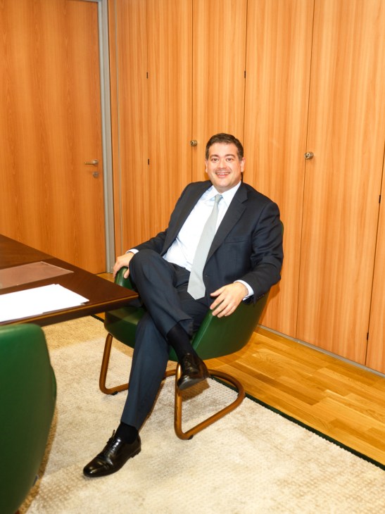 Man in dark suit and tie sitting in green chair against wooden wall paneling in office setting