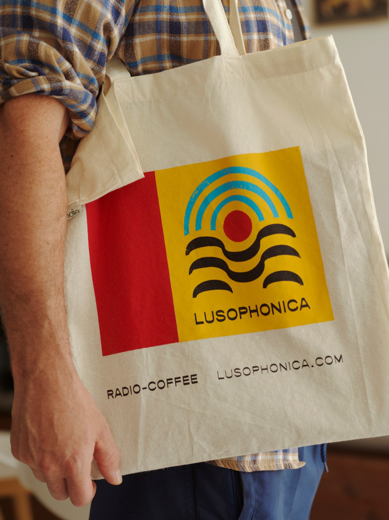 Person wearing plaid shirt carrying cream canvas tote bag with Lusophonica logo and website